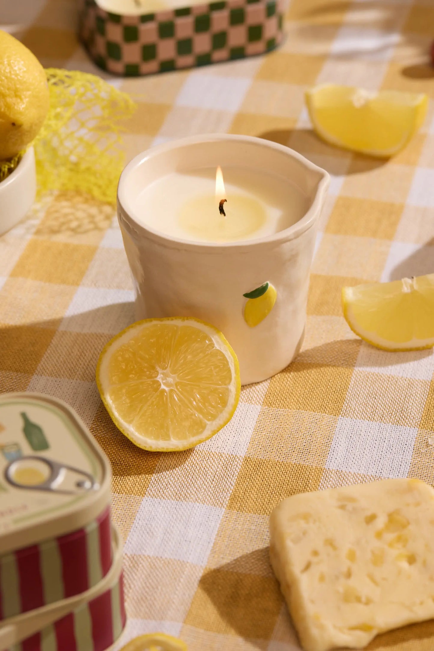 The Meyer Lemon - Bistro 8 oz Candle, with hand-painted food icons and a lemon design, sits on a yellow checkered tablecloth next to lemon slices, cheese, and a tin container.