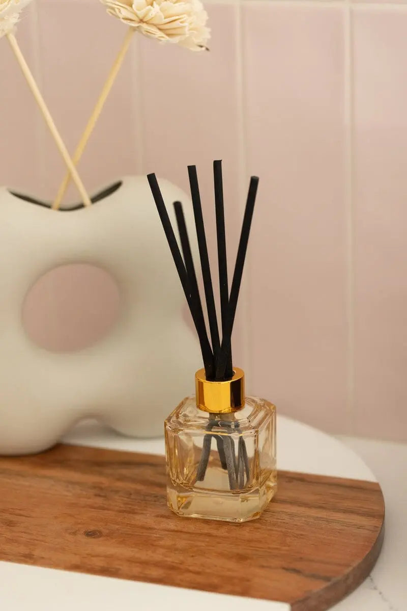 The Cotton + Teak - Botanica Diffuser 2.88 oz, featuring black reeds and a gold cap, is displayed on a wooden tray with a white vase and set against a pink tiled background.