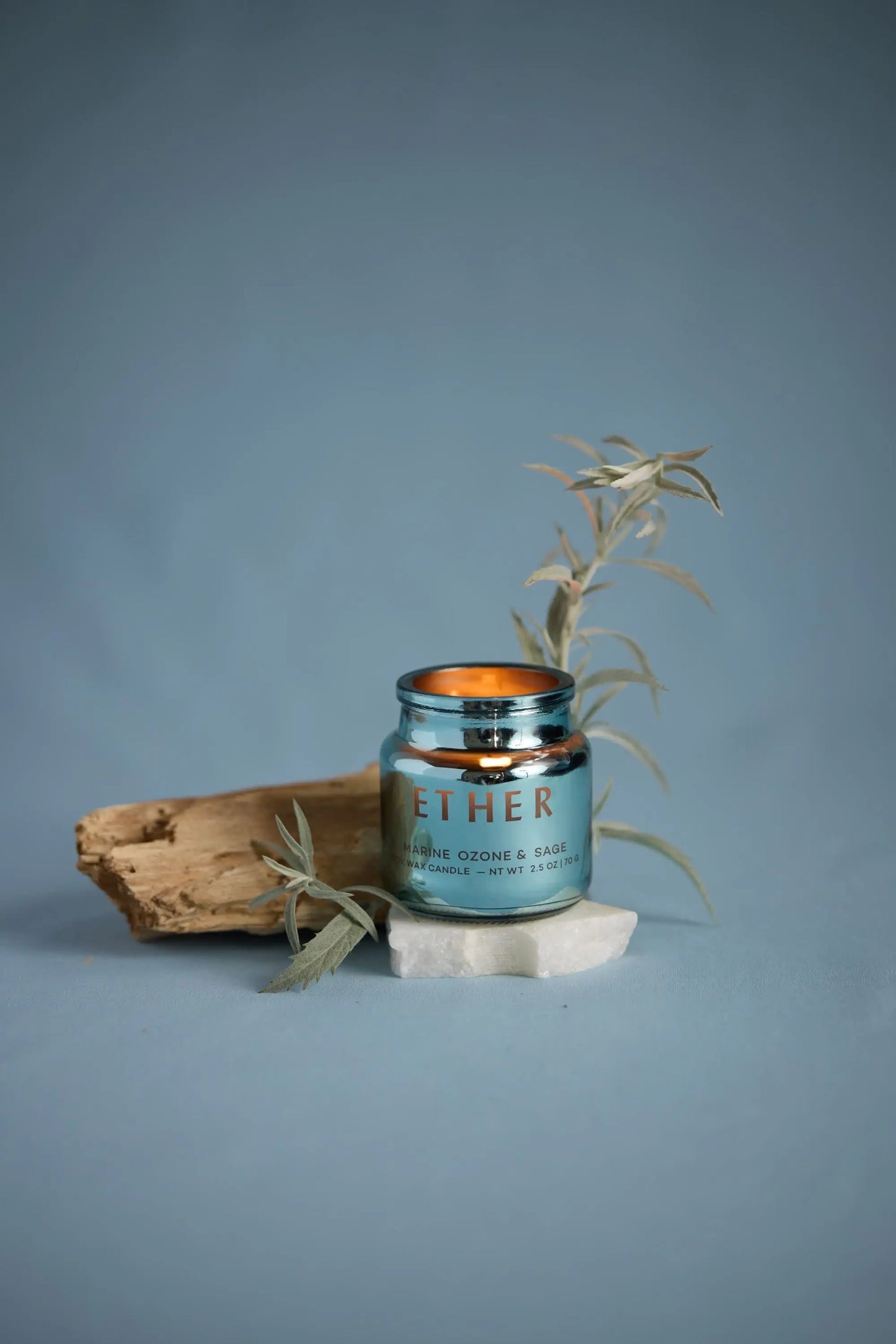 A blue Sea Salt & Sage - Chroma Candle 2.5 oz rests on a white stone, with a piece of wood and silvery foliage beside it, set against a blue background.