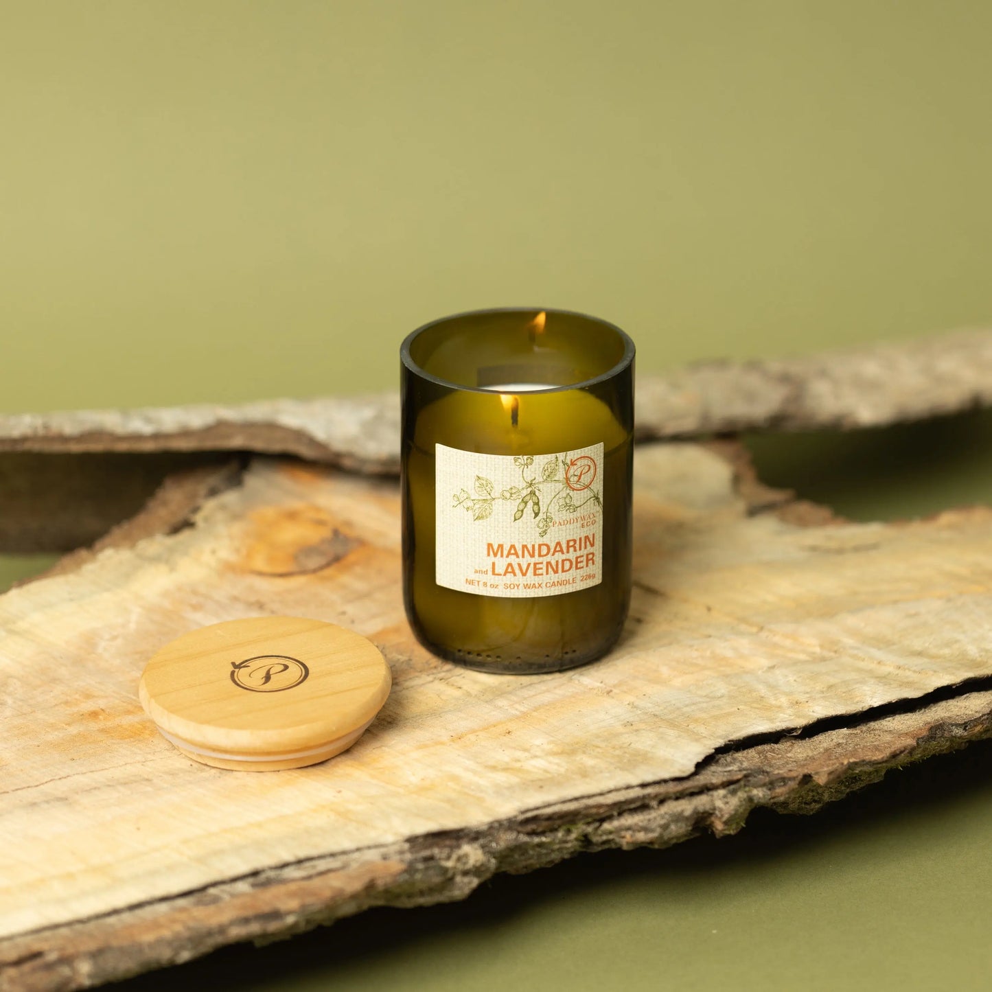 The Mandarin + Lavender - Eco 8 oz Candle, in a dark green glass jar with a round wooden lid, sits on wood against a green backgroundโan inviting accent for sustainable living.