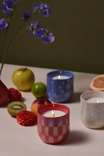 Three Cactus Flower - Lustre Candle 10 oz are lit in colored glass holders and arranged on a table with assorted fruits, flowers, and a green background.