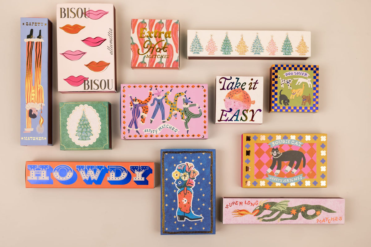 A collection of Cowboy Boot - Large Rectangle Boxed Matches featuring colorful illustrations of a cowboy boot, dogs, a cat, Christmas trees, lips, and a fire-breathing dragon.
