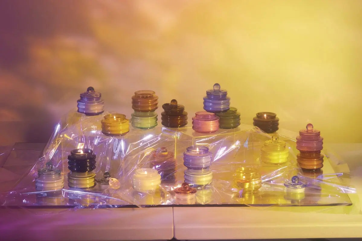 Stacked colorful glass discs and spheres sit on a reflective surface covered with clear plastic, next to the Lavender - Beam 3 oz Candle, all set against a yellow and purple gradient background.