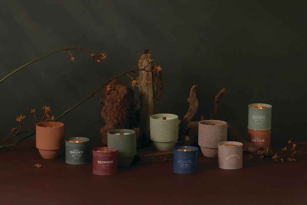 The Yellowstone - Parks 11 oz Candle, in a colored container with a crackling wood wick, sits on a dark surface and evokes a national park-inspired fragrance, surrounded by pieces of wood and dried branches.