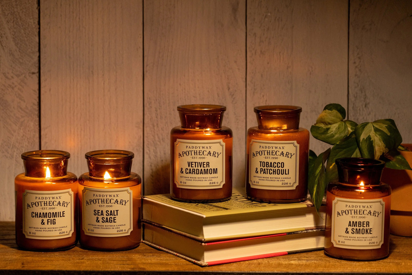 Three lit and two unlit Tobacco + Patchouli - Apothecary 8 oz candles in amber glass are arranged on stacked books and beside a plant, set against a wooden background.