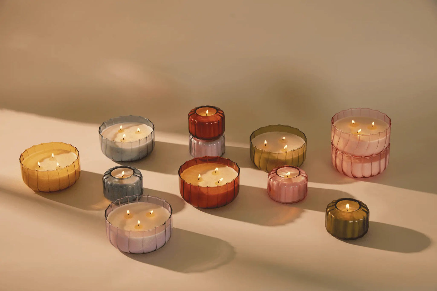 A lit Tobacco Patchouli - Ripple 4.5 oz Candle in a ribbed colored glass container is arranged on a neutral surface.