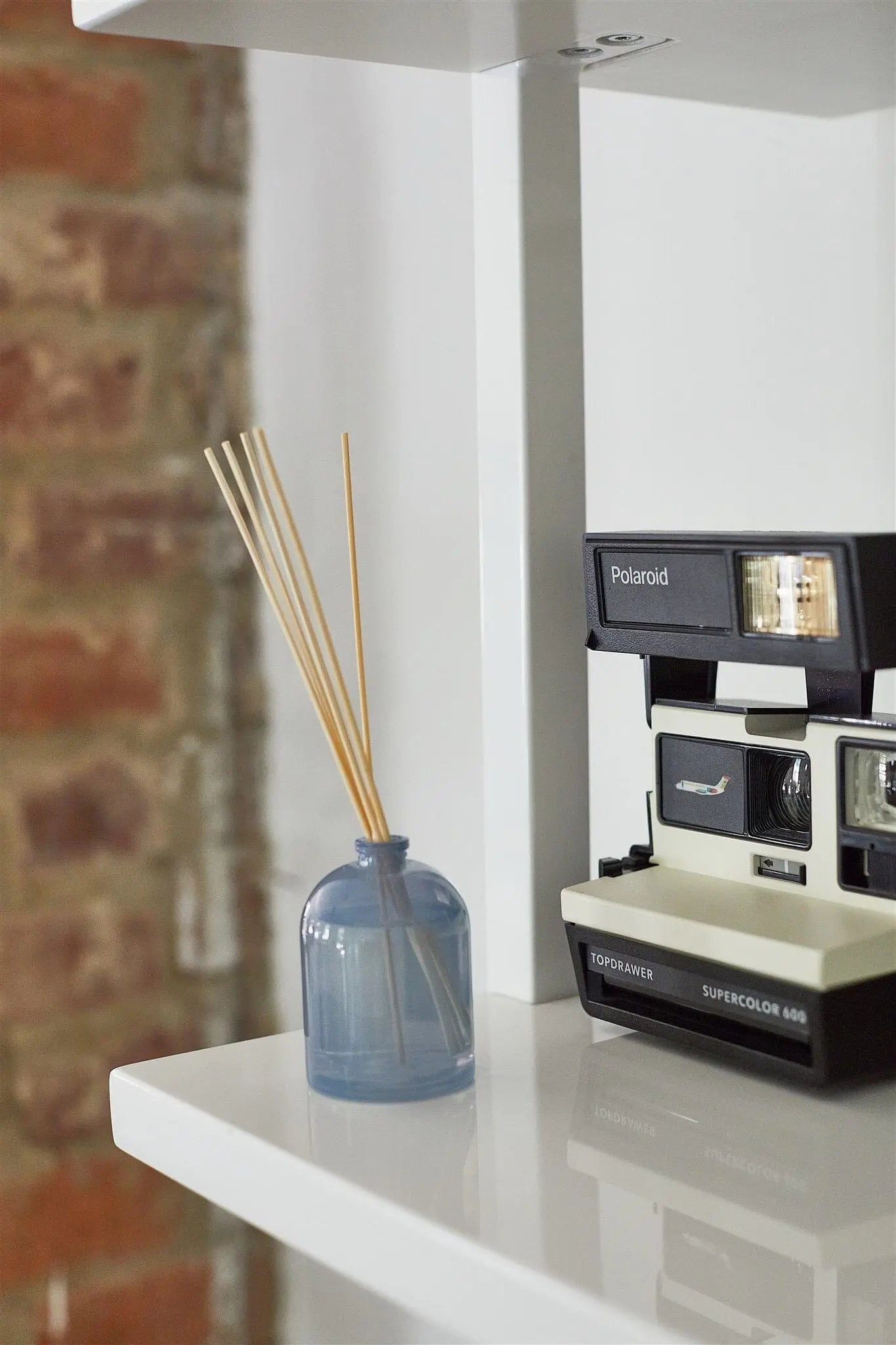 A Wild Fig - 1.5 oz Diffuser and a vintage Polaroid Supercolor 669 camera rest on a white shelf, creating a charming fragrance gift idea with a brick wall backdrop.