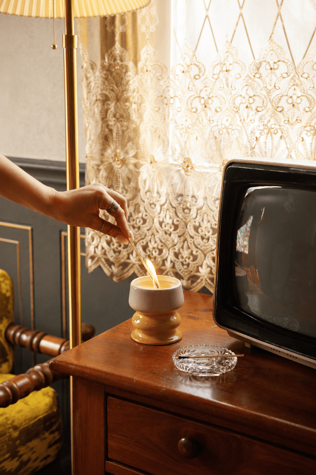 A hand lights the Smoked Pistachio - Melt 8.5 oz Candle on a wooden table next to an ashtray and an old television, with lace curtains and a vintage lamp in the background.