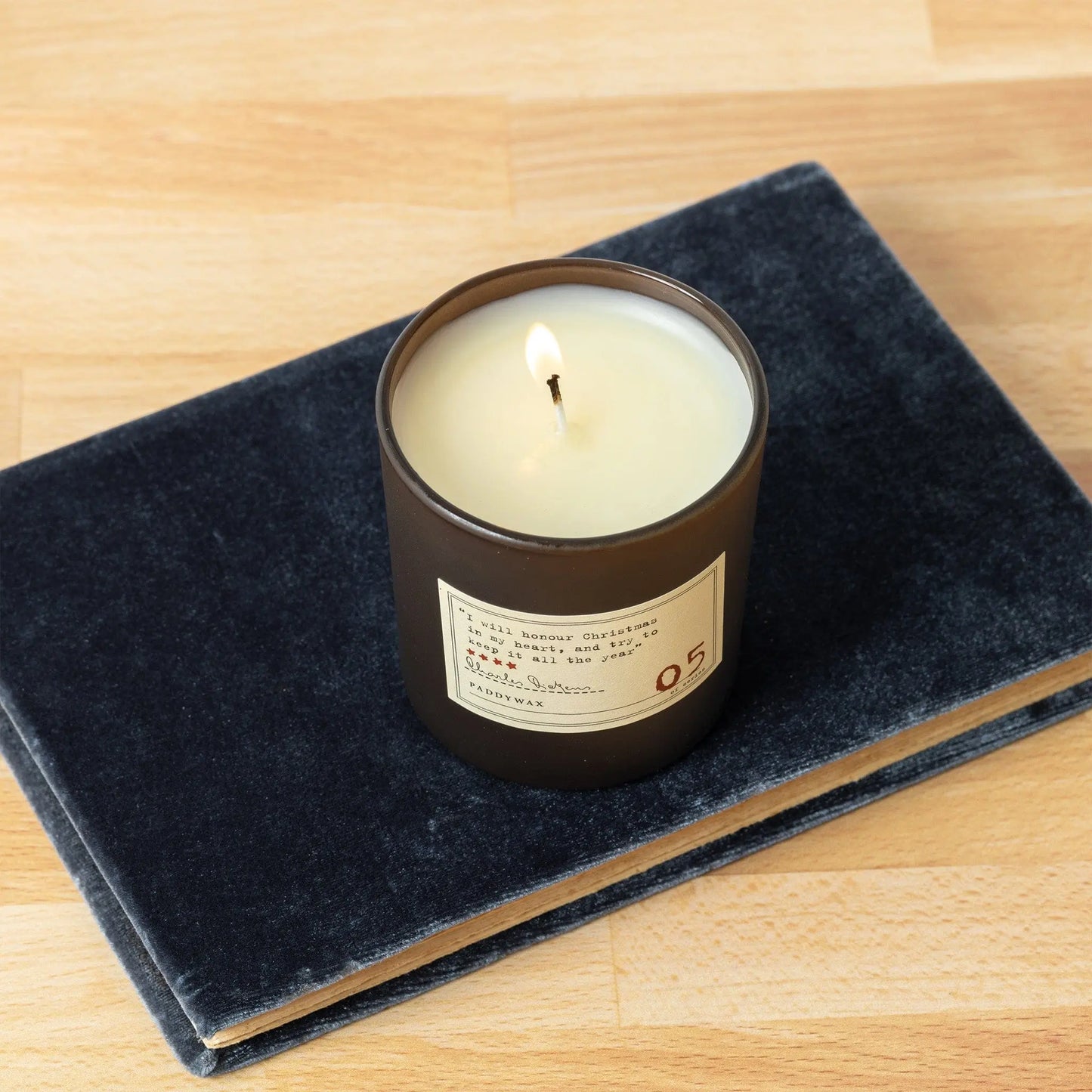 The Charles Dickens - Library Candle 6 oz from the Library Collection, in a dark glass container, rests on a closed dark gray book atop a light wood surface.