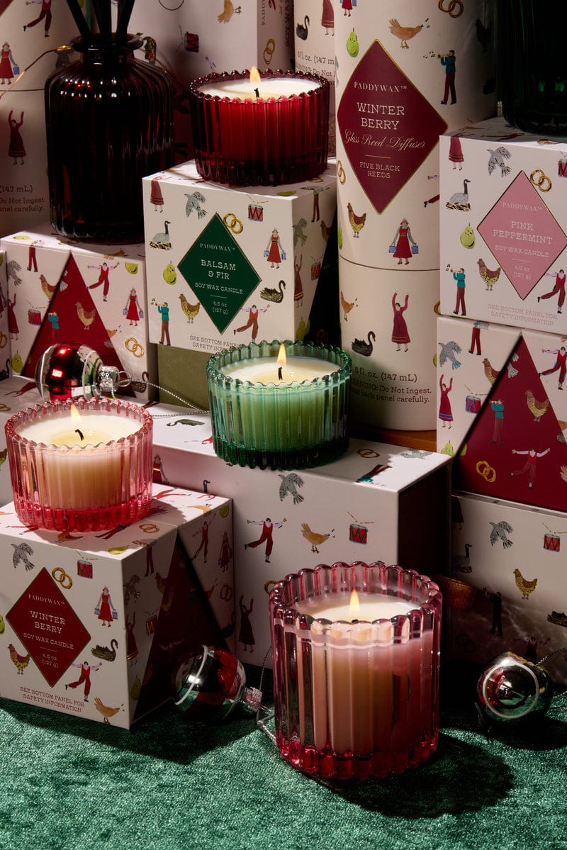 The Pink Peppermint - 12 Days Of Christmas candle glows in front of decorative boxes, spreading festive scents and adding to the holiday ambiance with ornaments and a green textured tablecloth.