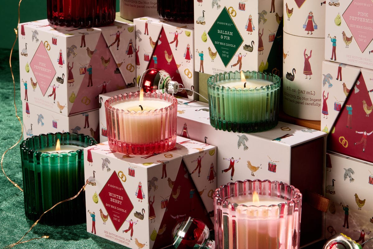 The Balsam & Fir - 12 Days Of Christmas features colorful ribbed glass candles in pink, green, and red atop festive illustrated holiday boxes, surrounded by ornaments and string lights for a cozy, celebratory scent.