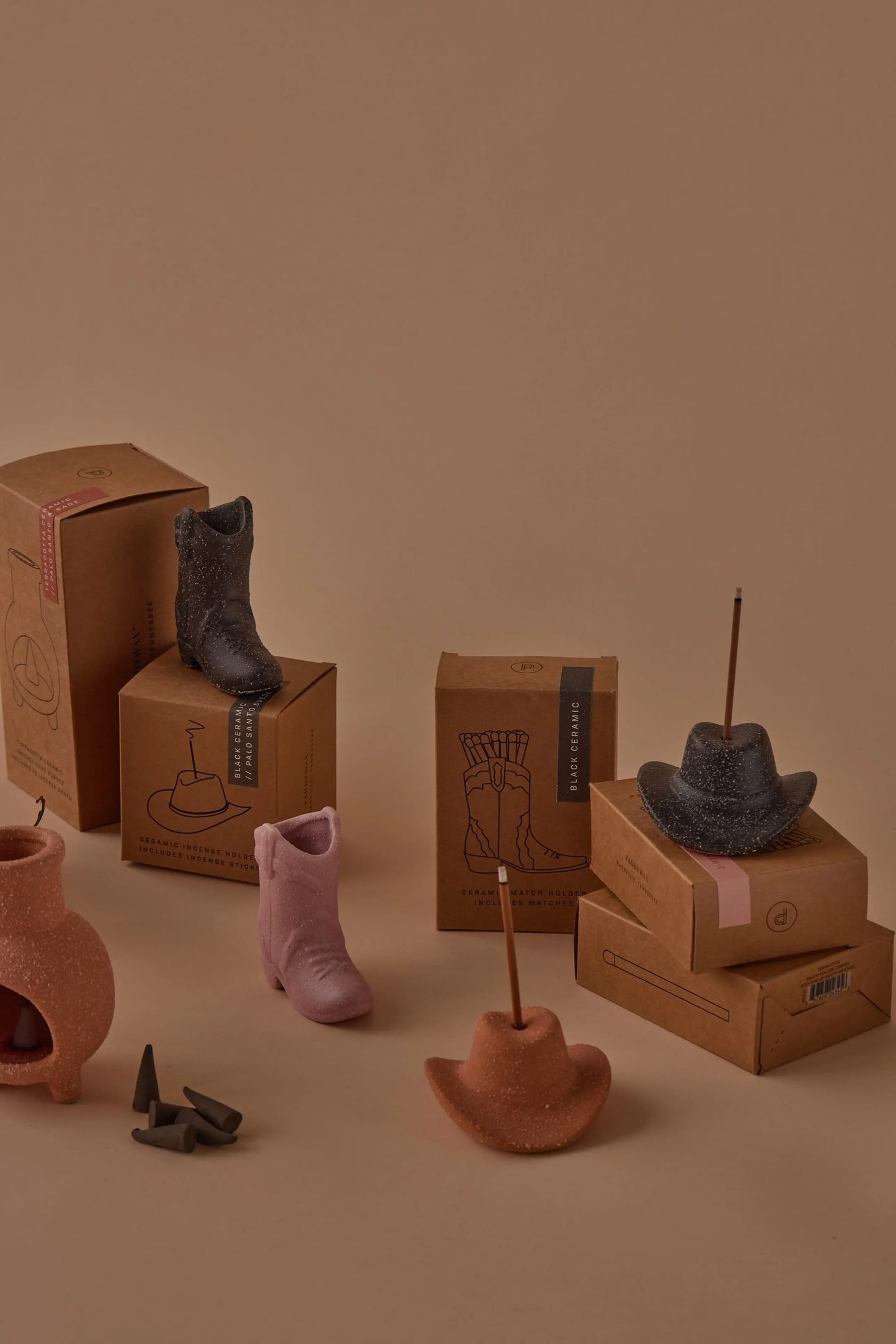 Teak & Tobacco - Incense Holders, including cowboy boots and hats, some with incense sticks, are arranged on and around cardboard boxes against a beige background.