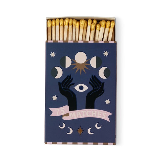 A Dopo "Moon" - 75ct Boxed Matches: Gold-tipped safety matches in a dark blue box with mystical designs—two black hands, an eye, moon phases, stars, and a pink "75 MATCHES" banner. Ideal for lighting your A Dopo candle or bringing Mediterranean vibes.