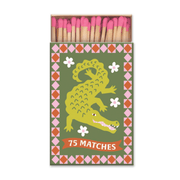 The A Dopo "Crocodile" - 75ct Boxed Matches feature an open matchbox with pink-tipped matches inside, a green crocodile with white flowers, a red-and-pink geometric border, and “75 MATCHES” in white at the bottom.