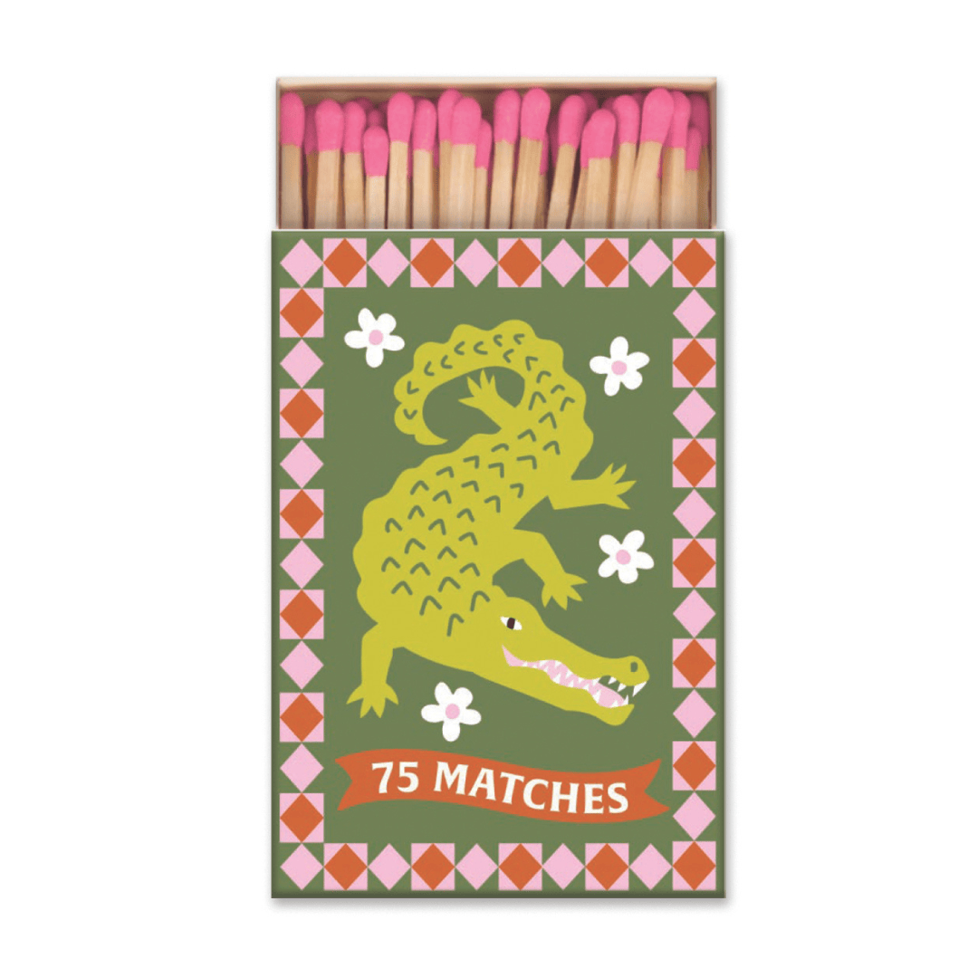 The A Dopo "Crocodile" - 75ct Boxed Matches feature an open matchbox with pink-tipped matches inside, a green crocodile with white flowers, a red-and-pink geometric border, and “75 MATCHES” in white at the bottom.