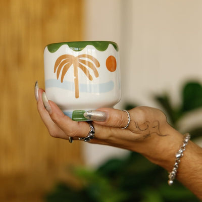 A hand with silver rings and long nails holds a mug decorated with a palm tree, green trim, and an orange sun, with blurred plants in the background.