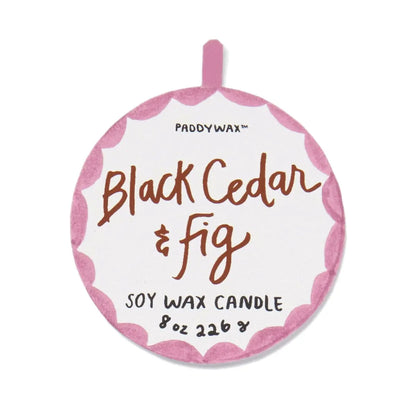 A round label with a pink scalloped border reads “Black Cedar & Fig - A Dopo 8 oz Candle” in handwritten-style text, evoking the allure of a Mediterranean fragrance.