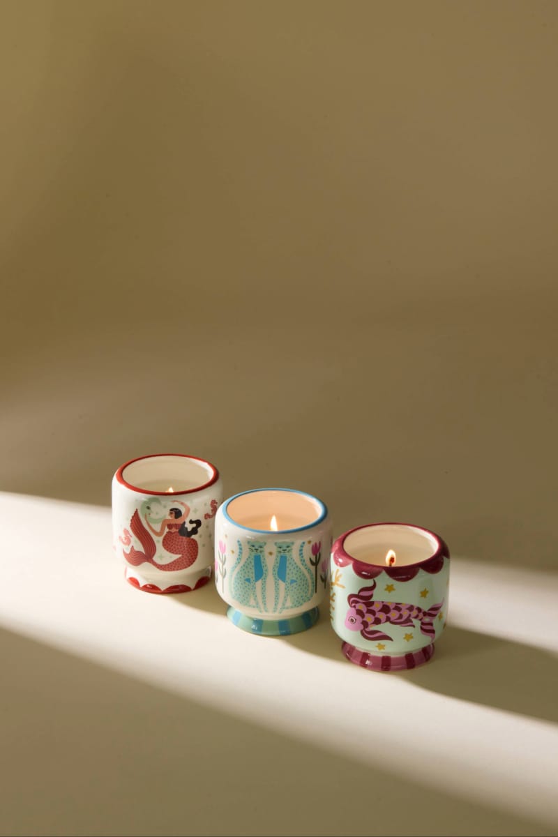 Three colorful, hand-painted ceramic candle holders, each cradling an A Dopo 8 oz Ginger & Teak candle with an earthy fragrance, are arranged in a row on a beige surface dappled with diagonal light and shadow.