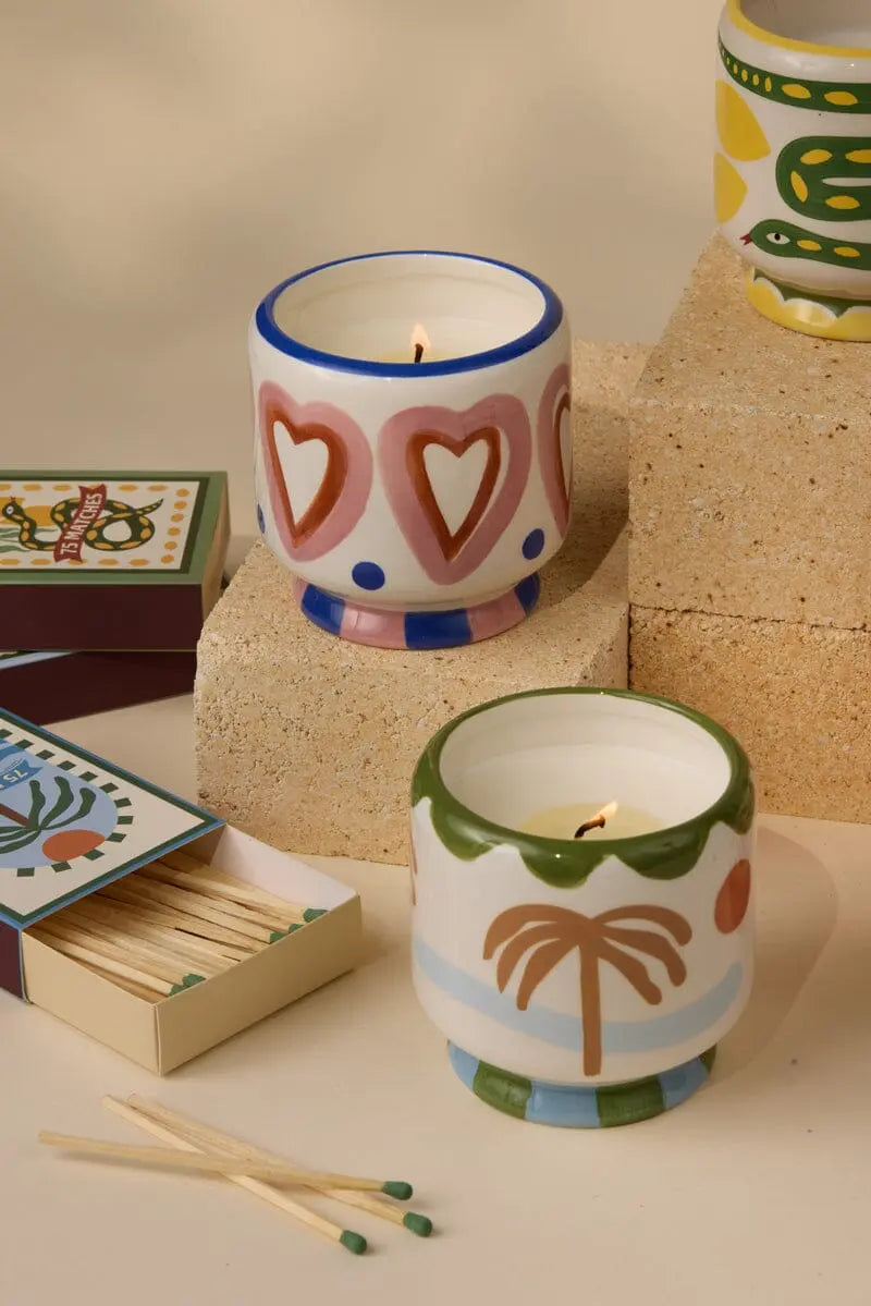 An 8 oz Rosewood Vanilla – A Dopo candle in a decorative holder rests on a textured block, surrounded by matchboxes and matches, filling the neutral setting with subtle Mediterranean-inspired fragrance.