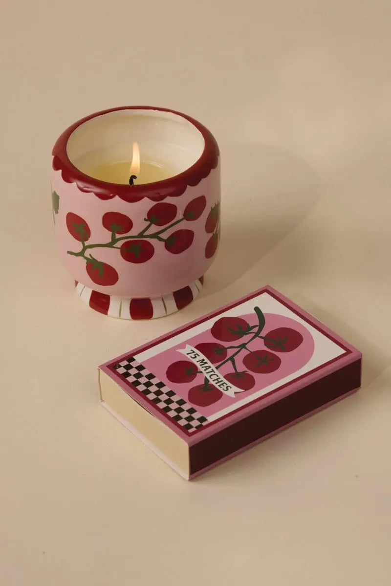 An A Dopo 8 oz Heirloom Tomato candle in a pink holder with red cherry designs sits beside its matching matchbox on a beige surface.