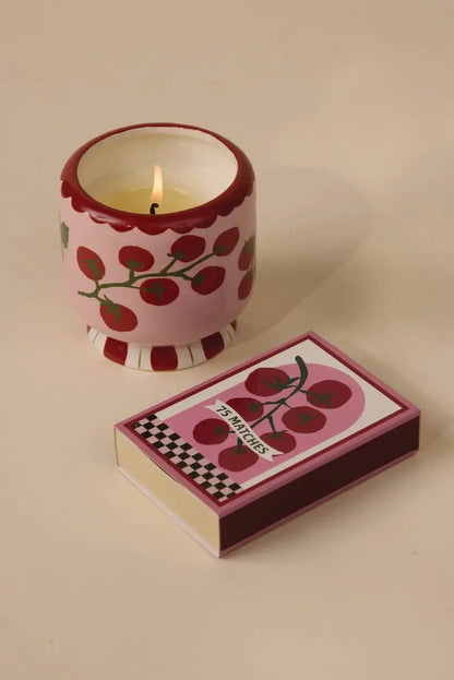 An A Dopo 8 oz Heirloom Tomato candle in a pink holder with red cherry designs sits beside its matching matchbox on a beige surface.