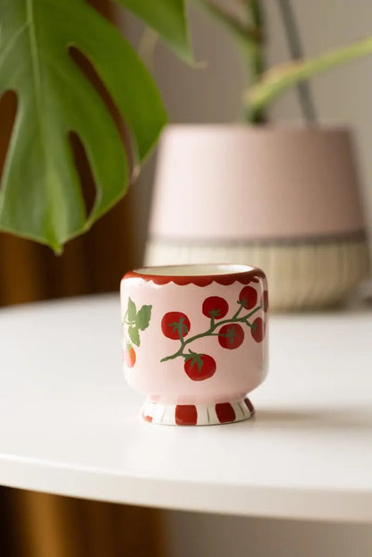 The Heirloom Tomato - A Dopo 8 oz Candle, in a small pink ceramic cup with a red tomato vine design, is perfect for candle lovers and displays beautifully on a white table with greenery in the background.