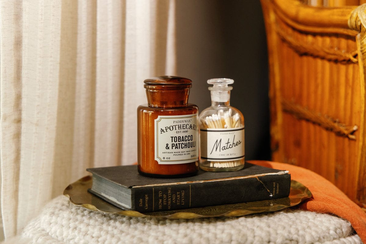 Two vintage apothecary bottles, one labeled Tobacco & Patchouli and the other Matches, sit on a tray atop a book, next to a wicker chair and a soft, textured surface in warm lighting.