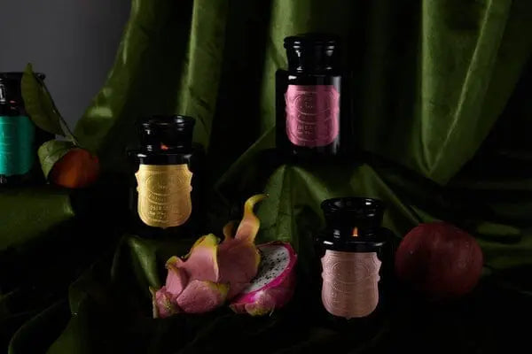 The Linen & Orris - Apothecary Noir 8 oz Candle in a dark glass jar with a colorful label is shown on green fabric, next to a halved dragon fruit and a whole peach.