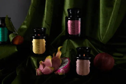 The Linen & Orris - Apothecary Noir 8 oz Candle in a dark glass jar with a colorful label is shown on green fabric, next to a halved dragon fruit and a whole peach.