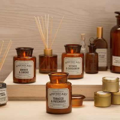 A display of amber glass jars and bottles labeled with various scents like Tobacco & Patchouli, Amber & Smoke, and Vetiver & Cardamom, alongside reed diffusers and small gold tins on wooden shelves.