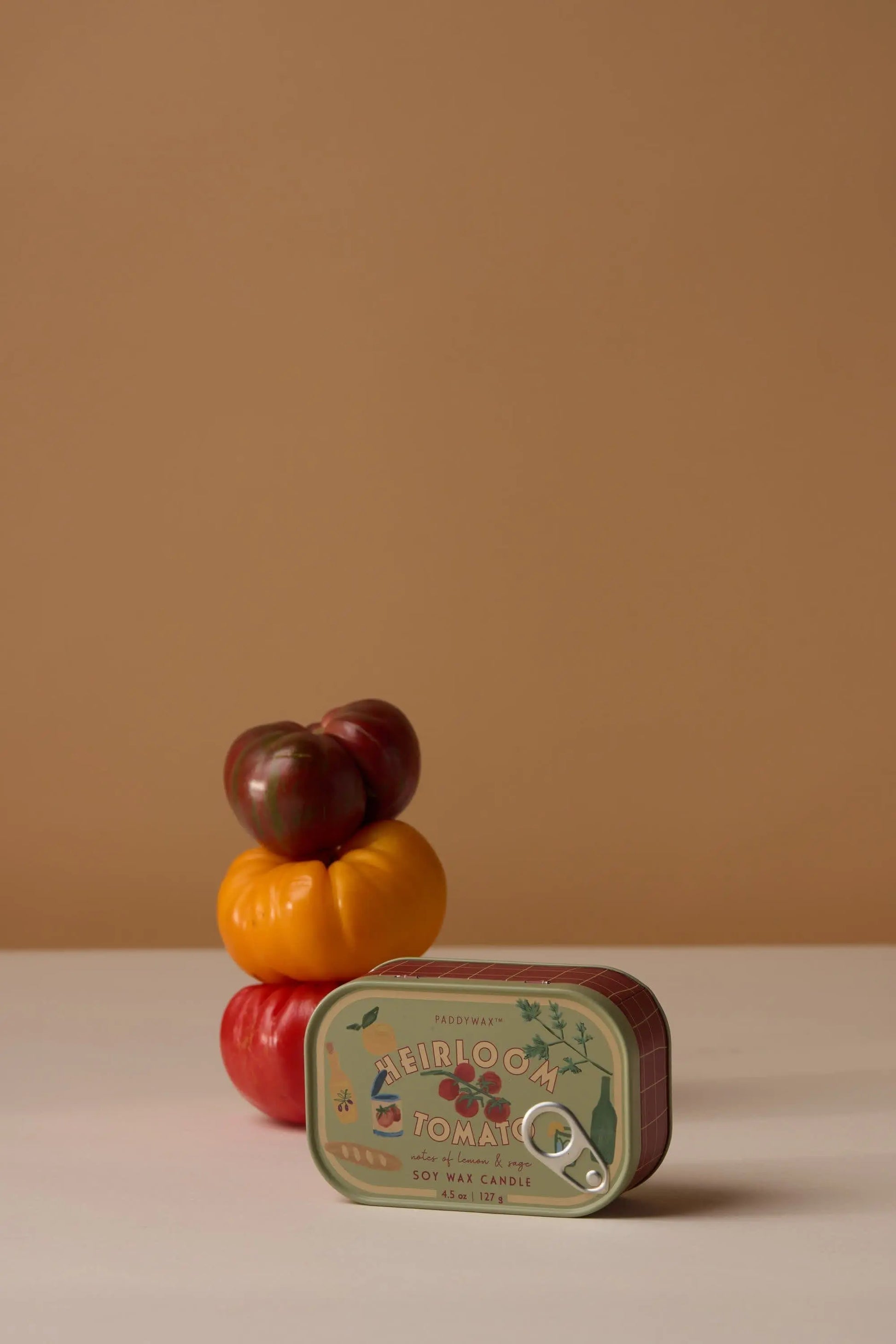 Three heirloom tomatoes—dark red, yellow, and red—are stacked on a white surface with the Heirloom Tomato - Bistro 4.5 oz Tin Candle in front, its label visible. The background is plain beige, evoking a subtle Parisian café ambiance.