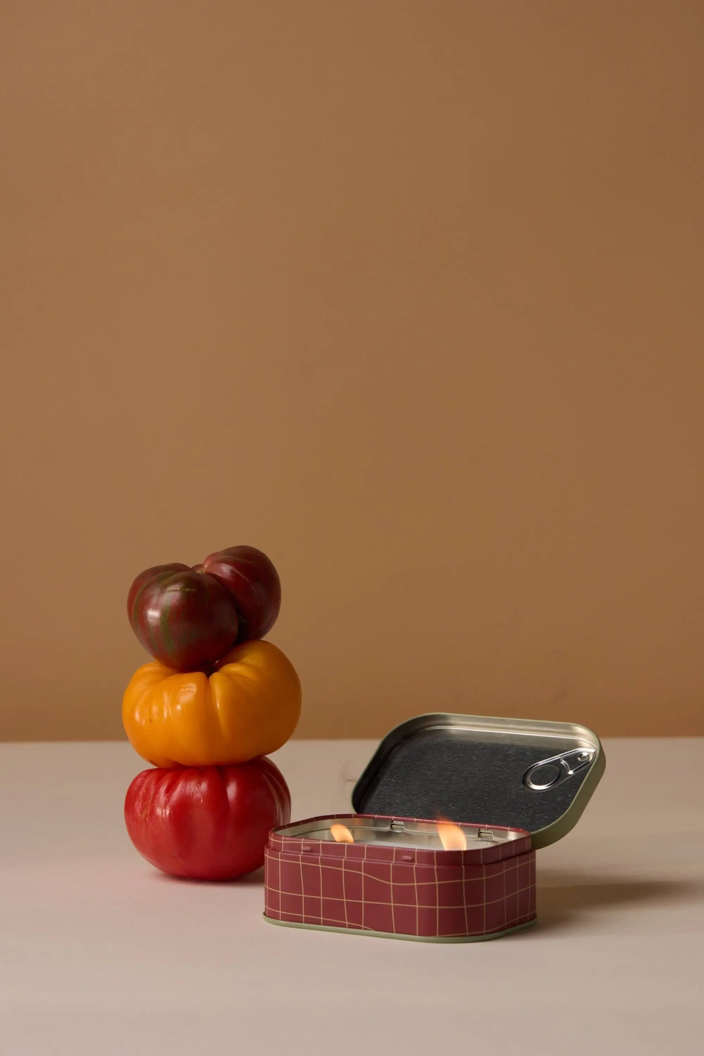 Three heirloom tomatoes—red, yellow, and dark red—are stacked beside an open tin. In front is the Heirloom Tomato - Bistro 4.5 oz Tin Candle with a red-checked design and small flame, all set against a plain beige background.