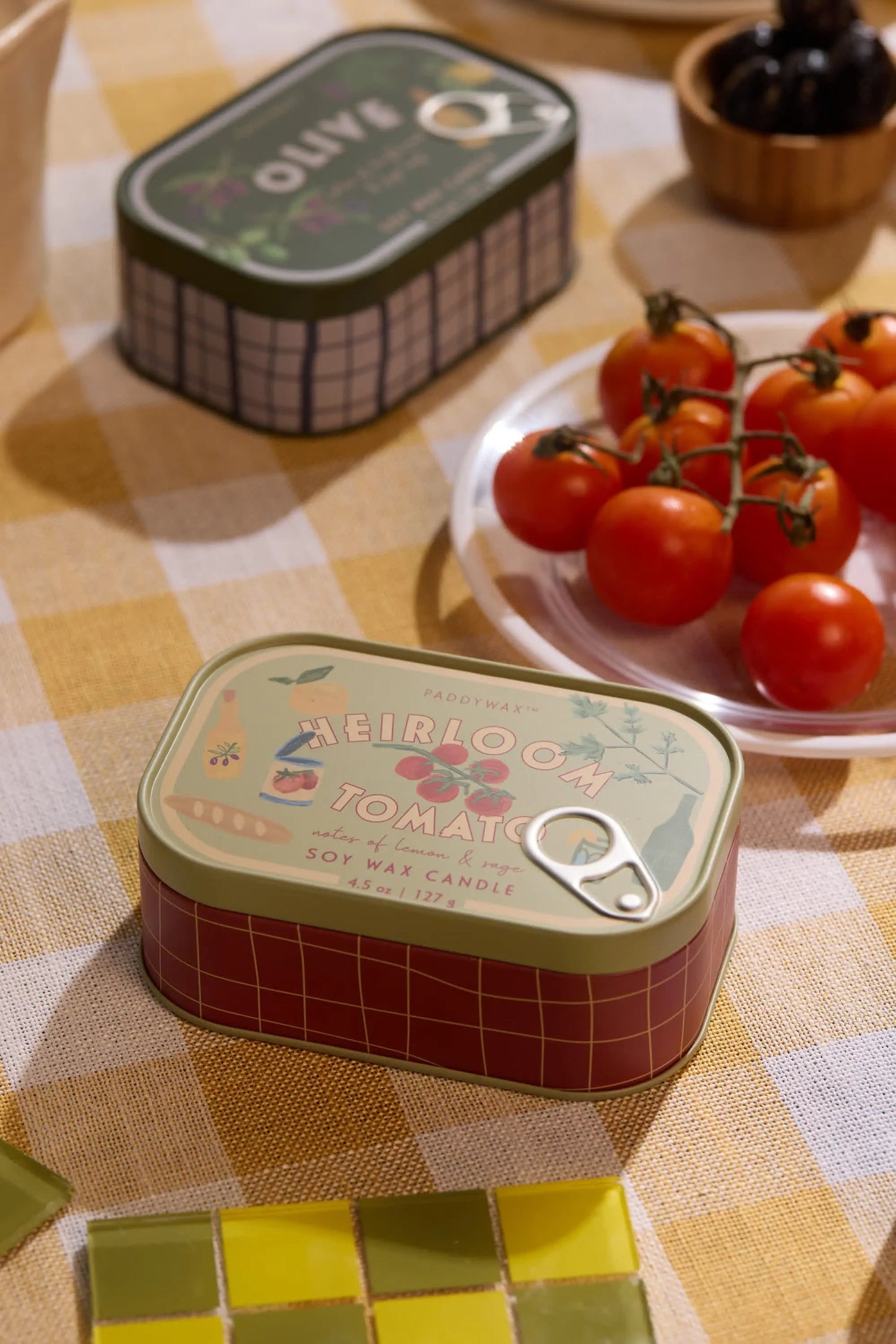 A Heirloom Tomato - Bistro 4.5 oz Tin Candle sits on a yellow plaid tablecloth beside a plate of cherry tomatoes, with another candle tin and small food bowls in the background.