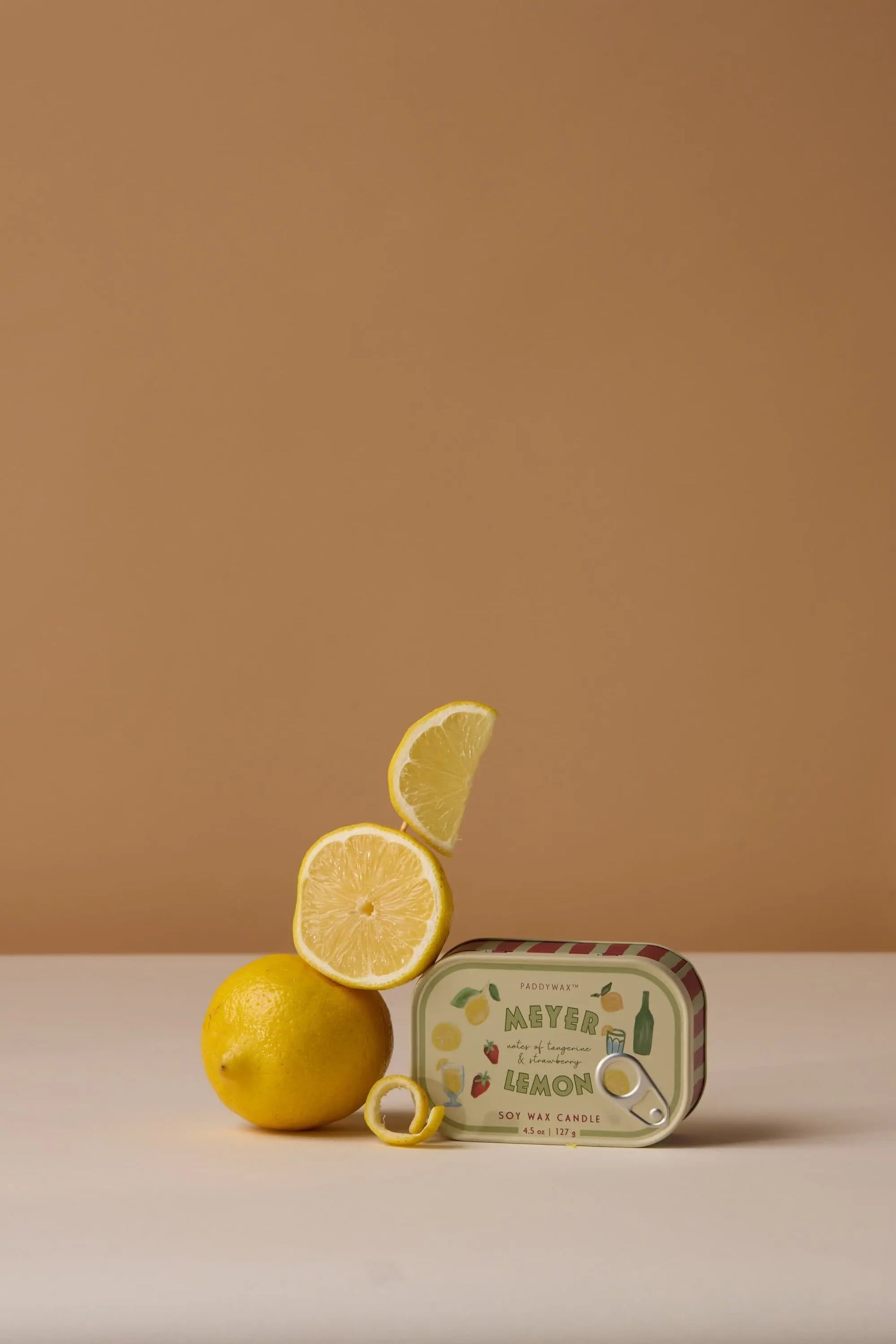 A whole lemon, lemon slice, and wedge are stacked beside the Meyer Lemon - Bistro 4.5 oz Tin Candle. A yellow ring sits in front of the candle, all arranged against a beige background.