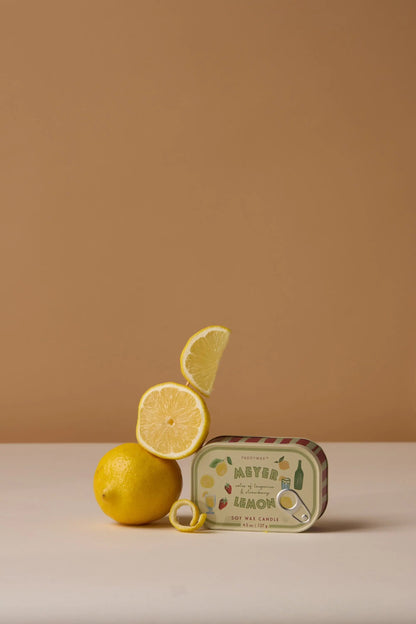 A whole lemon, lemon slice, and wedge are stacked beside the Meyer Lemon - Bistro 4.5 oz Tin Candle. A yellow ring sits in front of the candle, all arranged against a beige background.