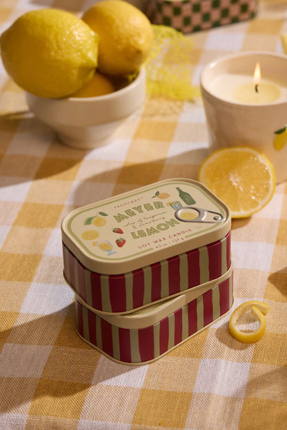 Two stacked Meyer Lemon - Bistro 4.5 oz Tin Candles rest on a yellow checkered tablecloth, surrounded by a sliced lemon, whole lemons in a bowl, another candle, and a yellow ribbon in warm light.