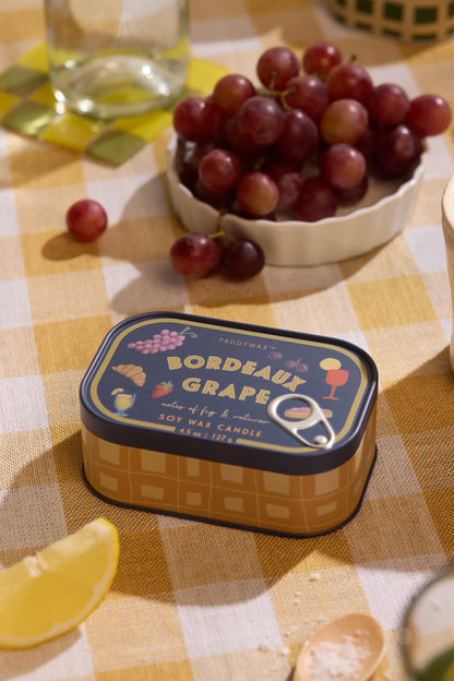 The Bordeaux Grape - Bistro 4.5 oz Tin Candle rests on a yellow checkered tablecloth beside red grapes, a lemon wedge, and a glass, evoking a cozy picnic atmosphere.