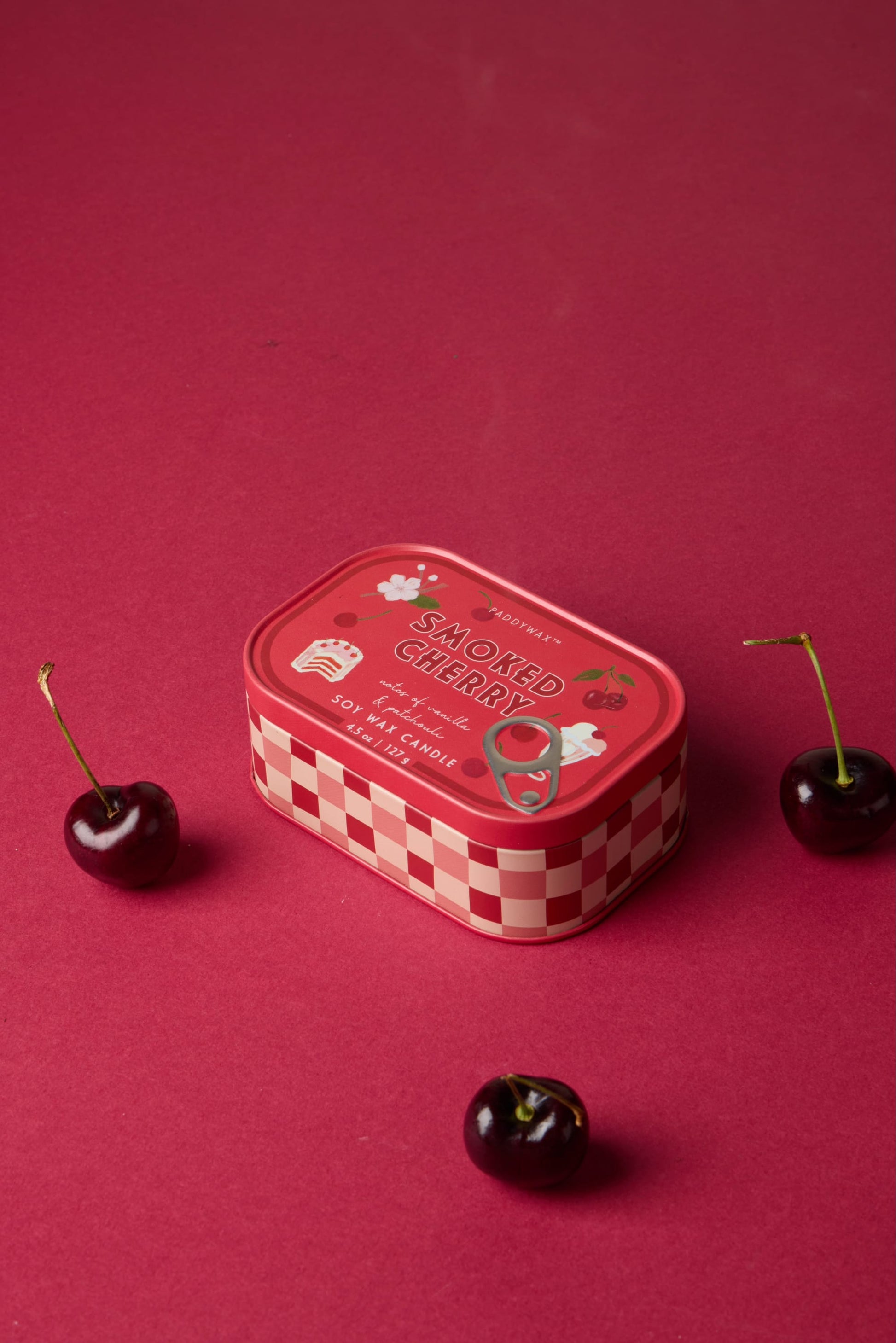 A Smoked Cherry - Bistro 4.5 oz Tin Candle with a red checkered border and illustrated cherries on the lid rests on a red surface, surrounded by three fresh cherries.