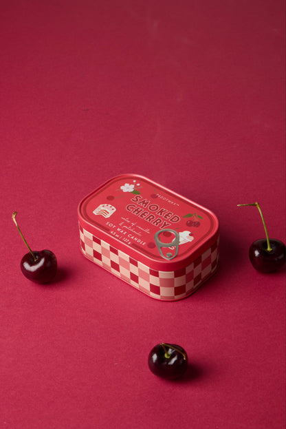 A Smoked Cherry - Bistro 4.5 oz Tin Candle with a red checkered border and illustrated cherries on the lid rests on a red surface, surrounded by three fresh cherries.