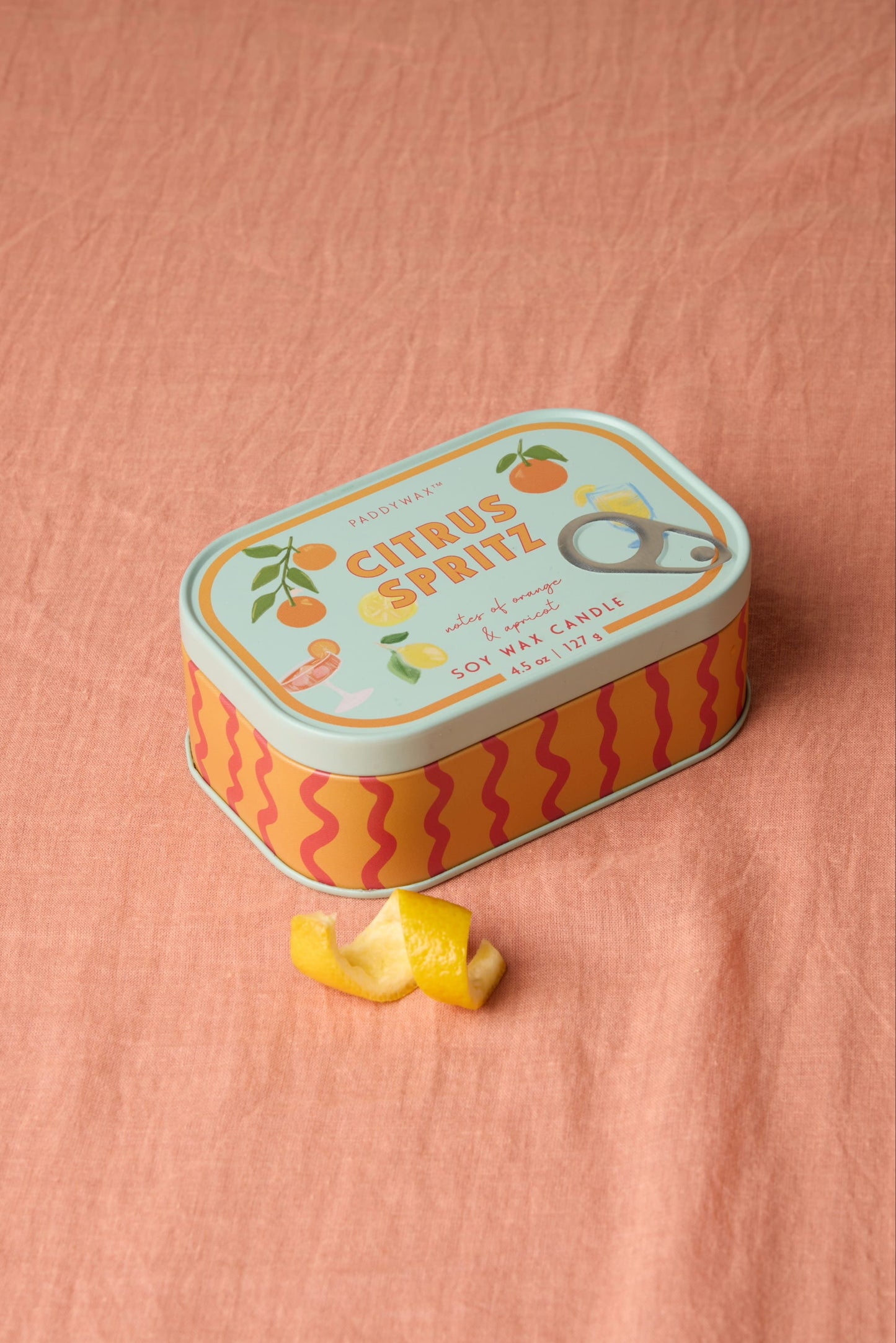 The Citrus Spritz - Bistro 4.5 oz Tin Candle, featuring orange designs on a sardine-style tin, rests on pink fabric with a curled lemon peel in front.