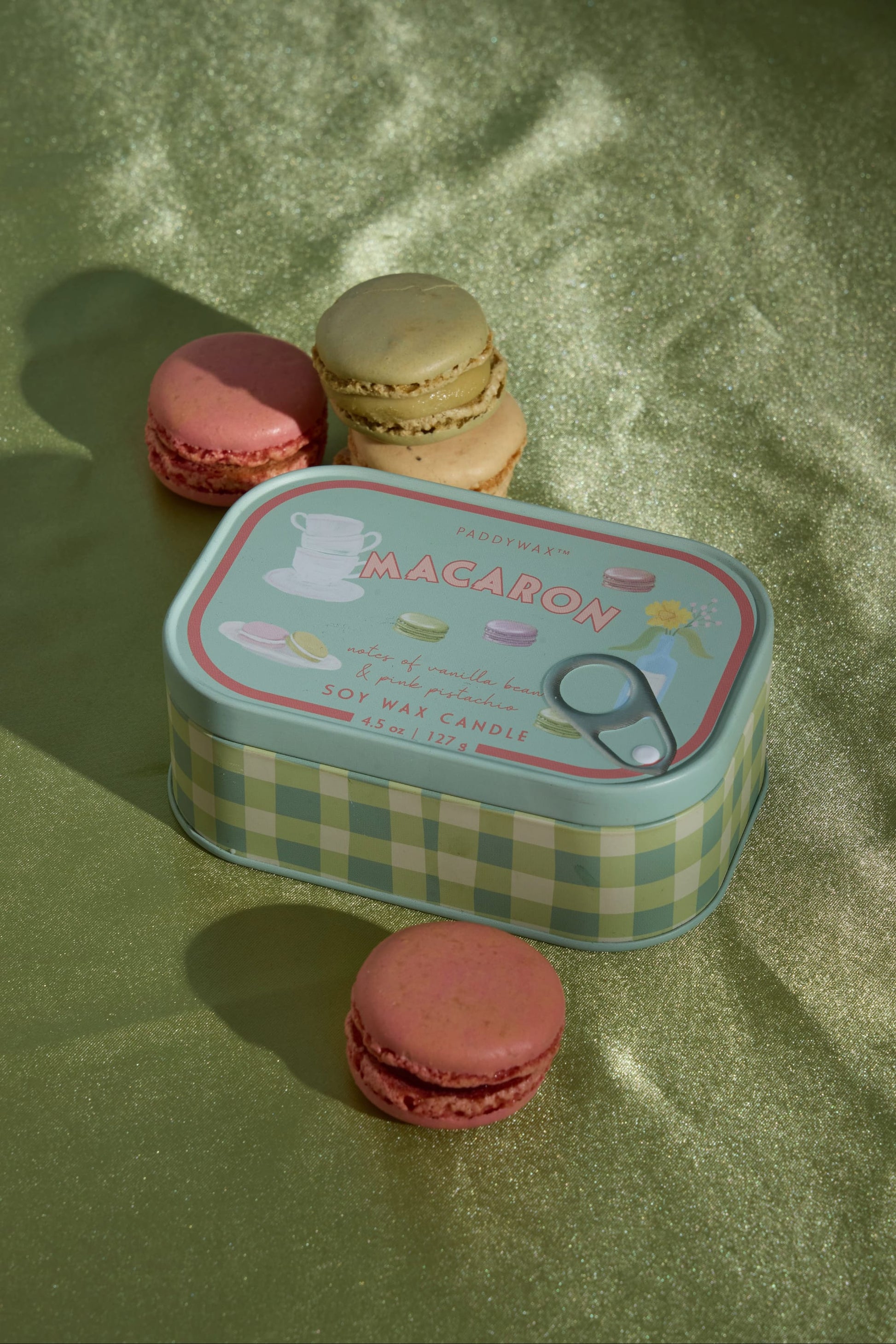 The Macaron - Bistro 4.5 oz Tin Candle rests on a green surface, surrounded by four vibrant pink and yellow macarons. Infused with a sweet macaron scent, this French-inspired candle evokes the delightful ambiance of a Parisian patisserie.