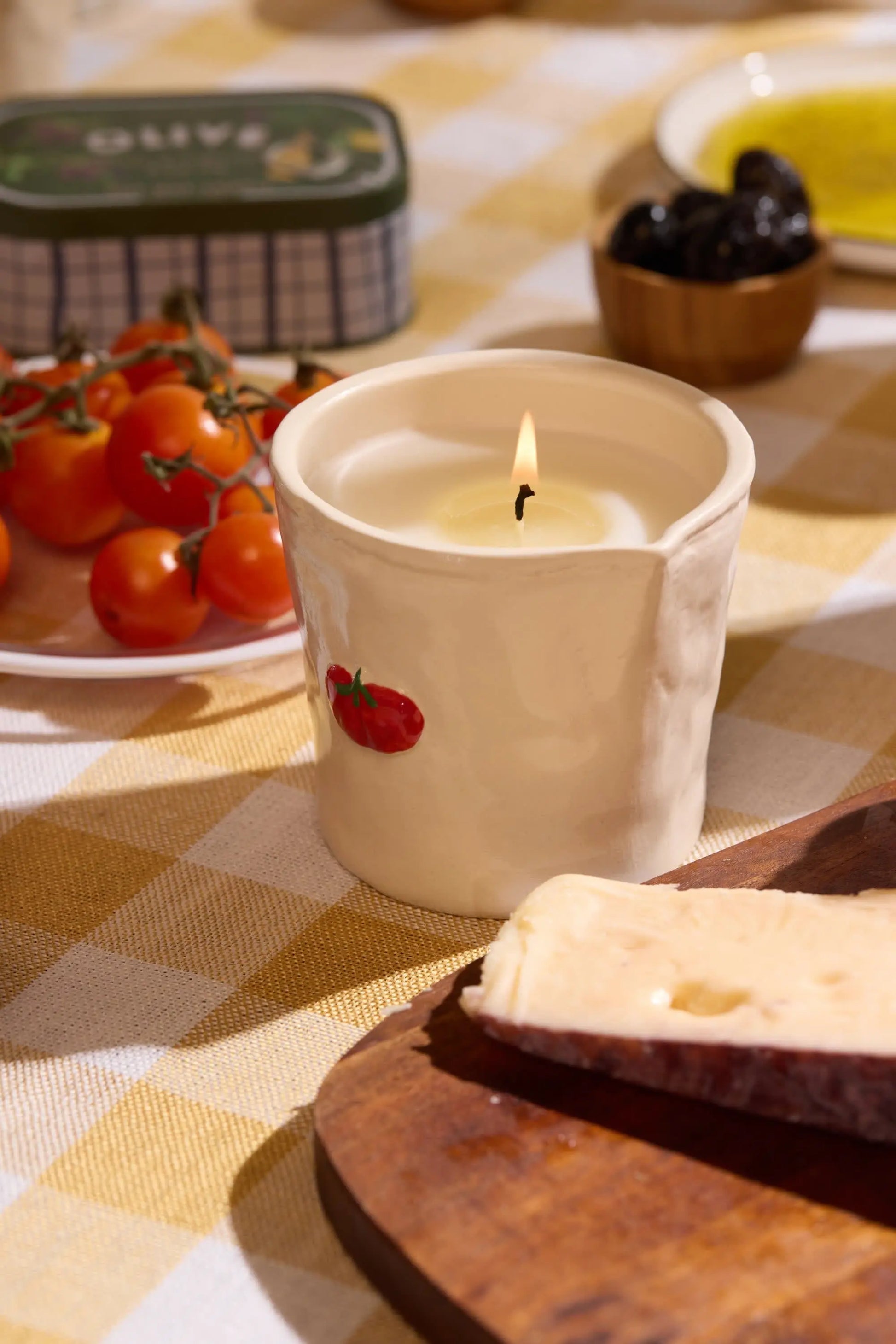 The Heirloom Tomato - Bistro 8 oz Candle, in a white holder with a tomato design, is lit on a checkered tablecloth near cherry tomatoes, cheese on a wooden board, and other food items.
