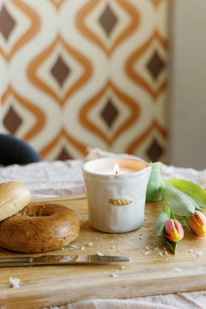 The Baguette - Bistro 8 oz Candle in a white ceramic holder sits with a bagel, knife, and tulips on a wooden board, set against retro orange and brown geometric wallpaper.