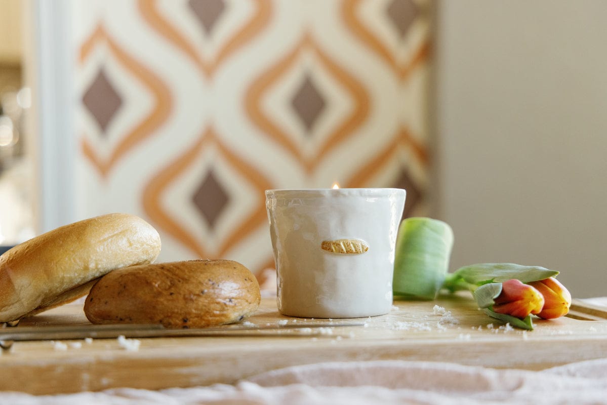 Candle on a wooden surface with bread and flowers, against a patterned wall.