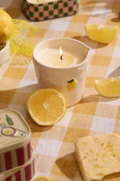 The Meyer Lemon - Bistro 8 oz Candle, with hand-painted food icons and a lemon design, sits on a yellow checkered tablecloth next to lemon slices, cheese, and a tin container.