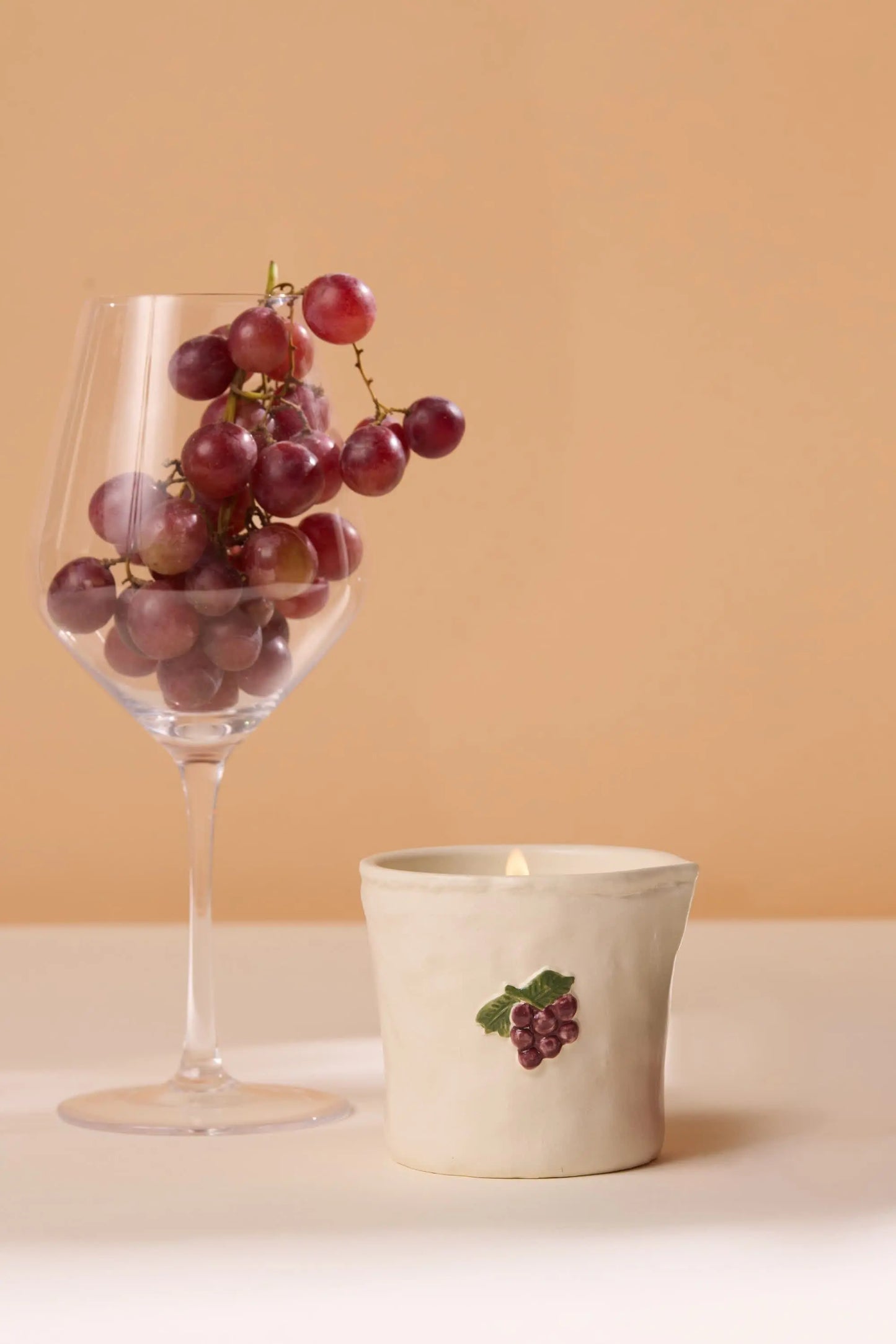 A wine glass of red grapes stands beside a white candle holder with a grape motif, set against beige—evoking cozy Parisian cafés with the Bordeaux Grape - Bistro 8 oz Candle’s aromatic fragrance and ceramic charm.