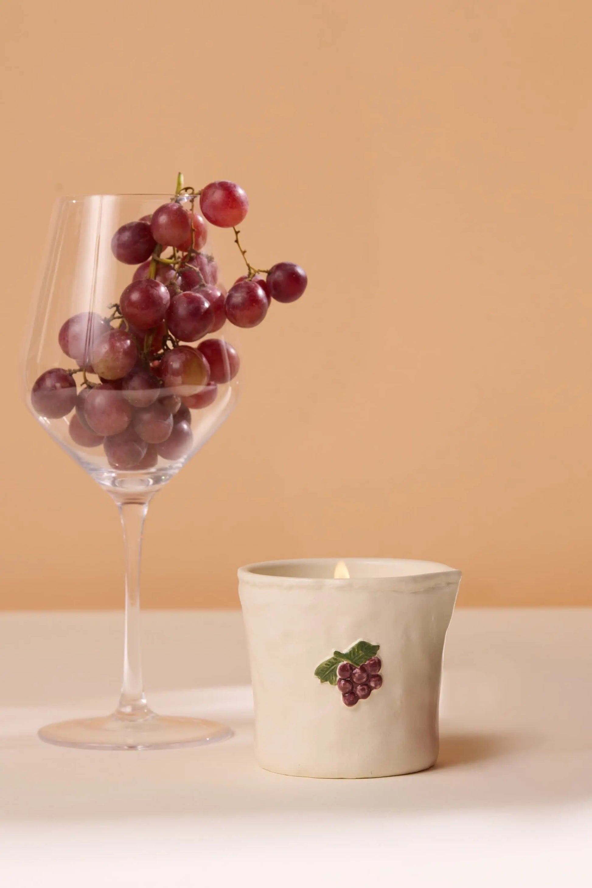 A wine glass of red grapes stands beside a white candle holder with a grape motif, set against beige—evoking cozy Parisian cafés with the Bordeaux Grape - Bistro 8 oz Candle’s aromatic fragrance and ceramic charm.