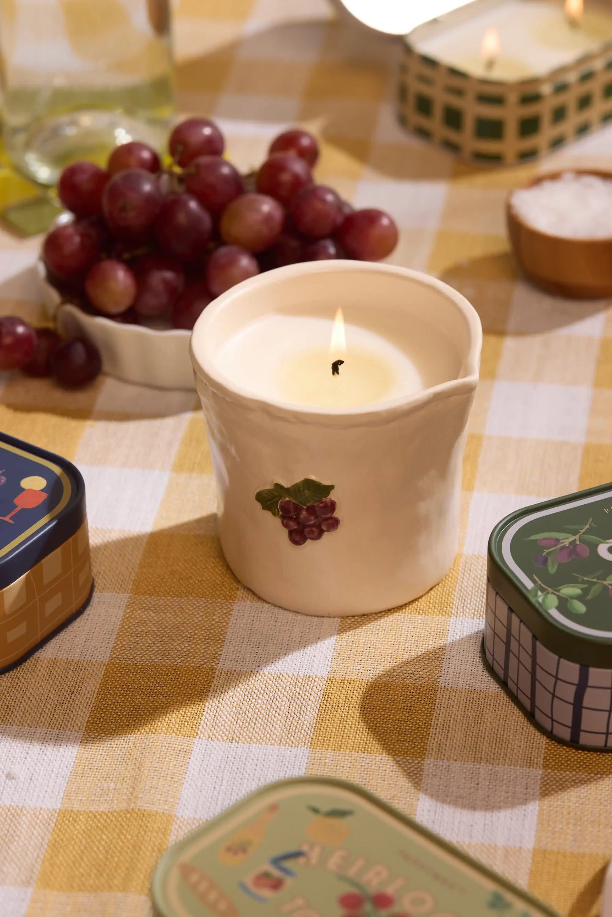 The Bordeaux Grape - Bistro 8 oz Candle, in a grape-themed ceramic holder, sits on a yellow checkered cloth—filling the air with Parisian café fragrance amid tins, grapes, and a small salt dish.