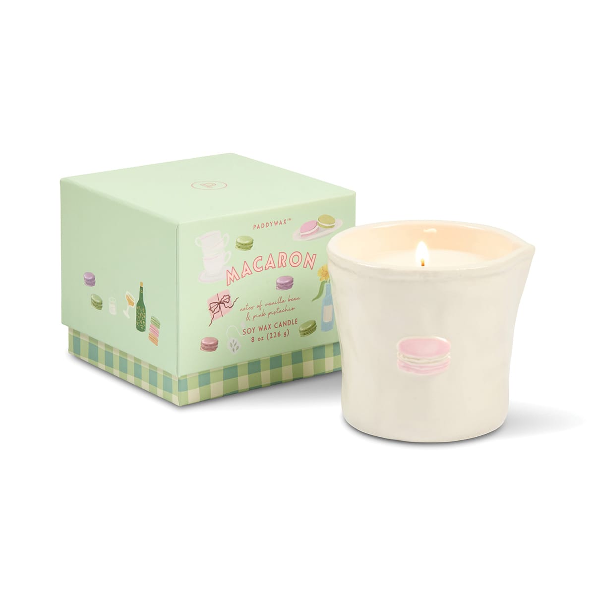 A lit Macaron - Bistro 8 oz Candle in a white, food safe ceramic holder adorned with a pink macaron illustration is displayed next to a pastel green box decorated with macarons and labeled “Macaron.” This charming setup blends delightful design with practical, food safe ceramics.