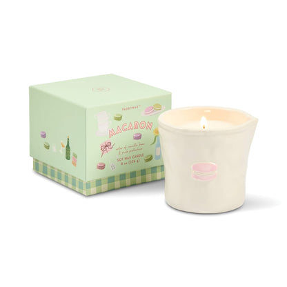 A lit Macaron - Bistro 8 oz Candle in a white, food safe ceramic holder adorned with a pink macaron illustration is displayed next to a pastel green box decorated with macarons and labeled “Macaron.” This charming setup blends delightful design with practical, food safe ceramics.